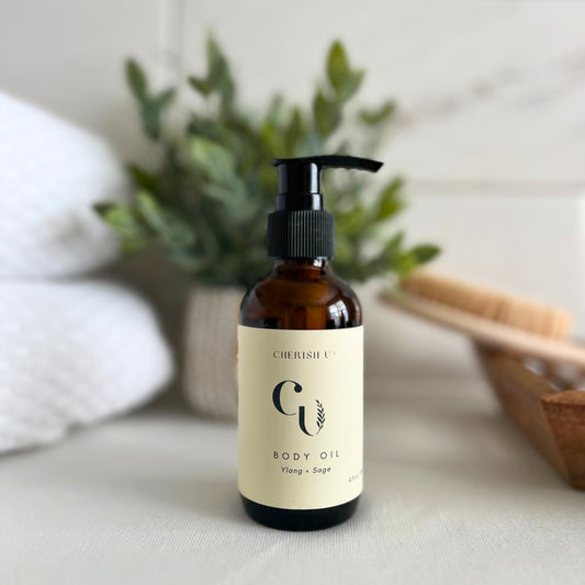 An amber bottle of Ylang + Sage body oil with a pump, beautifully arranged on counter with  greenery and fresh towels Cherish U®