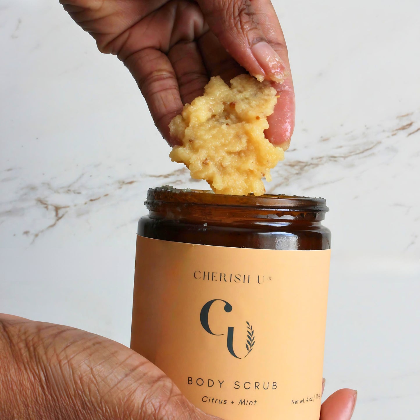 A hand holding a scoop of Citrus + Mint Sugar Scrub taken from an amber jar, showcasing the invigorating citrus and mint-infused exfoliant. Cherish U®