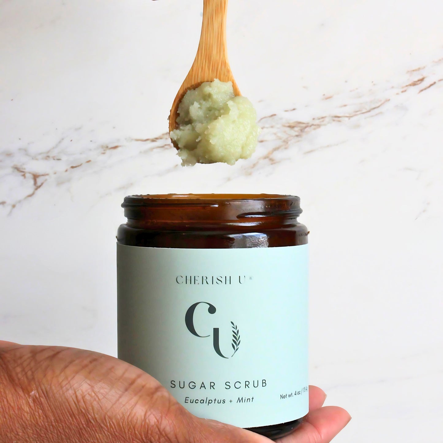 A wooden spoon scooping Eucalyptus + Mint Sugar Scrub from an amber jar, displaying the refreshing eucalyptus and mint-infused scrub for a rejuvenating exfoliation. Cherish U®