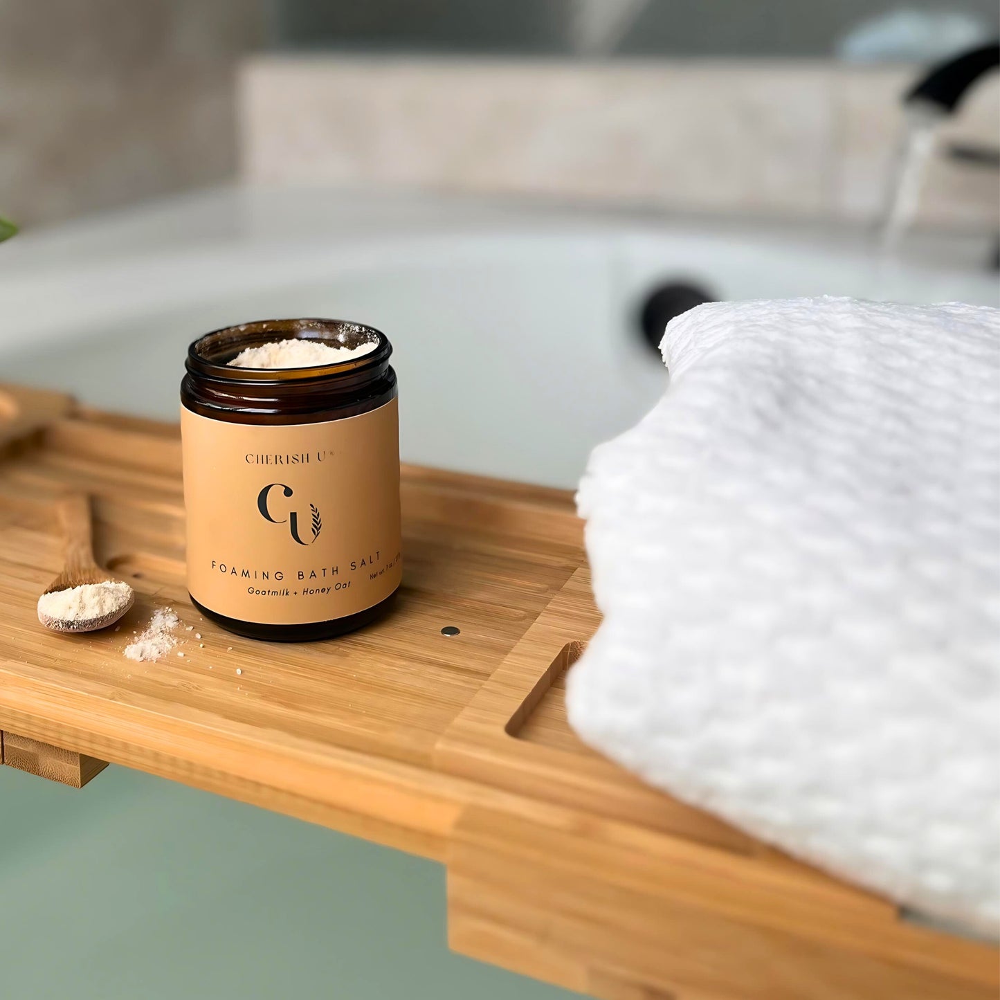 A jar of Goatmilk + Honey Oat foaming bath salts rests attractively on a wooden bathtub tray. The background features running bath water, highlighting a soothing and nourishing bath experience Cherish U®