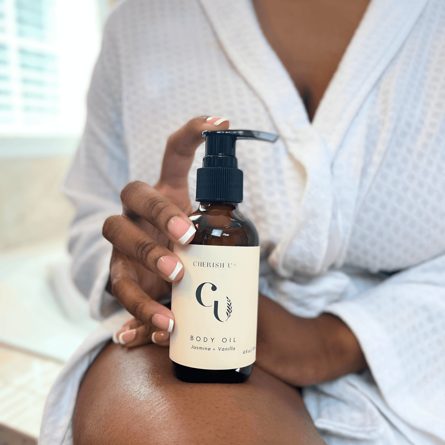 A person holding an amber bottle of Jasmine Vanilla body oil with a practical pump, seated on the edge of a bathtub in a bathroom. Their skin glows with a freshly oiled sheen, highlighting the luxurious and fragrant experience Cherish U®