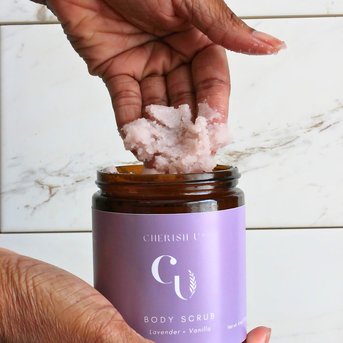 A hand holding a portion of Lavender + Vanilla Sugar Scrub scooped from an amber jar, highlighting the calming lavender and warm vanilla blend for gentle exfoliation. Cherish U®