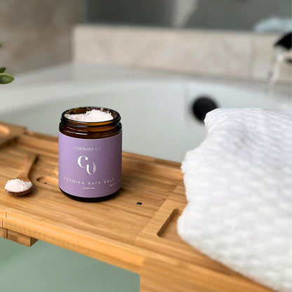 A jar of Lavender foaming bath salts sits elegantly on a wooden bathtub tray. In the background, water flows into the tub, creating a relaxing and inviting atmosphere