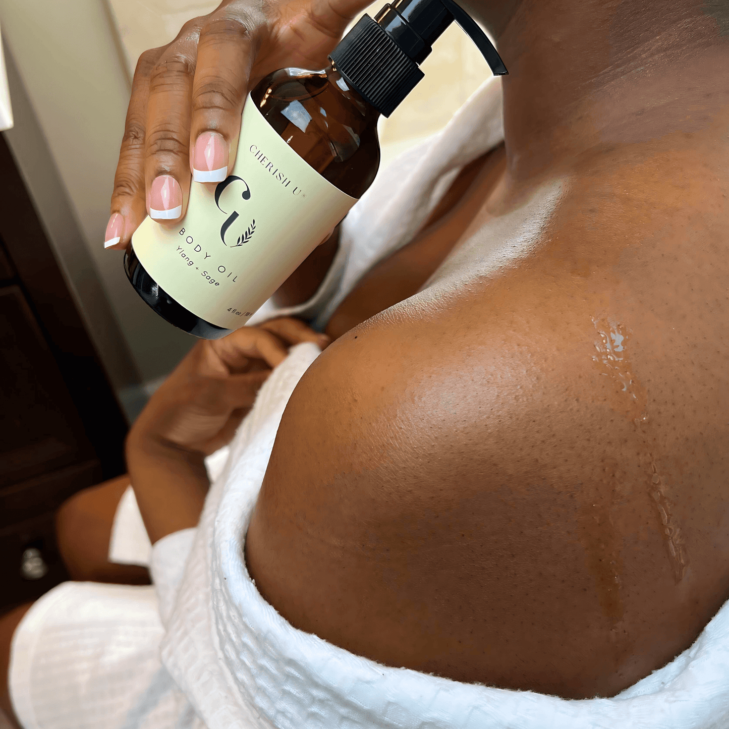 A person dispensing Ylang + Sage body oil from an amber bottle with a pump, rubbing the oil onto their shoulders. Their skin  glows from the freshly oiled sheen. Cherish U®